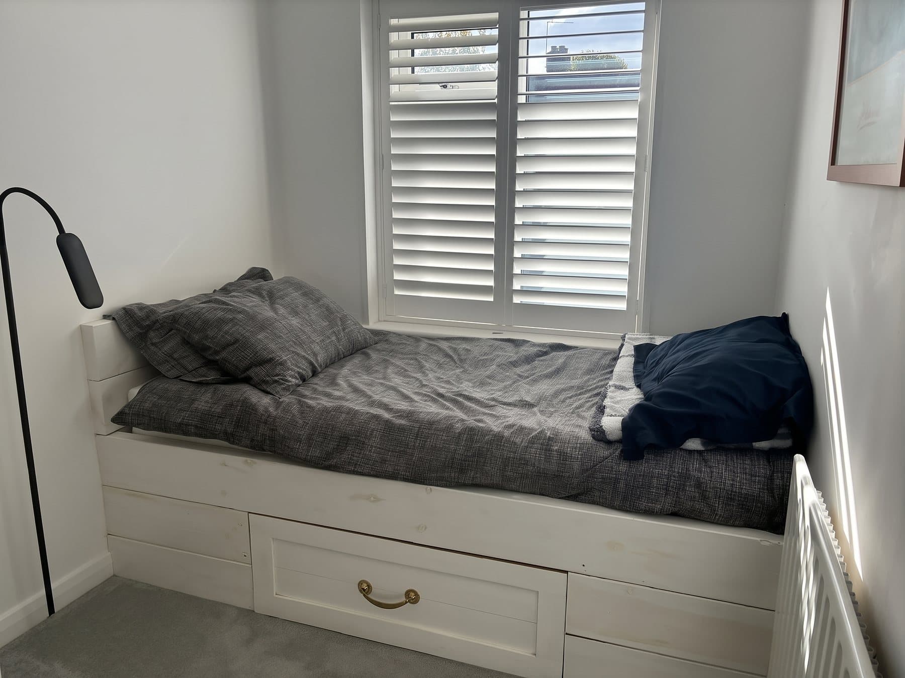 A single bed with a white wooden frame and storage drawers beneath it, under a window with white shutters.